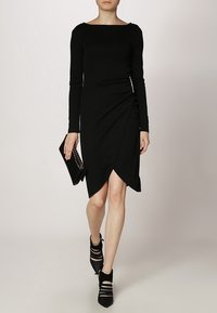 Černé dlouhé šaty s lodním výstřihem, s bočním překryvem a asymetrickým lemem. Oblečené s černou kabelkou a sandály na vysokém podpatku.