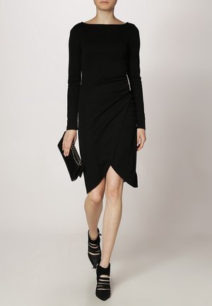 Musta pitkähihainen mekko venepääntiellä, jossa on sivulle kietoutuva muotoilu ja epäsymmetrinen helma. Yhdistetty mustan clutch-laukun ja remmikenkien kanssa.