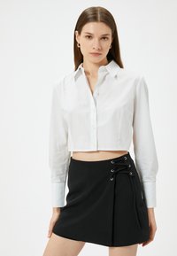 Camicia bianca a maniche lunghe, tagliata, con bottoni, abbinata a una gonna nera con dettagli di lacci sui lati, realizzata in un tessuto morbido.