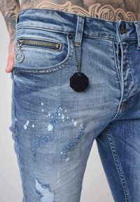 Light blue denim jeans with distressed details, a zip pocket, and a black octagonal tag attached by a chain. Smooth texture and casual design.