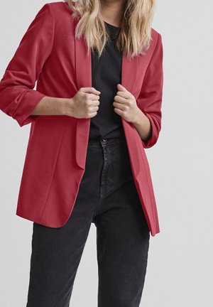 Person wearing a red blazer over a black shirt and black jeans, holding the blazer lapels with both hands.