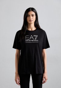 Melna kokvilnas T-krekls ar metāliski sudraba logotipu "EA7 EMPORIO ARMANI" uz priekšu. Īsas piedurknes un vaļīga piegriezums. Vienkārša tekstūra.