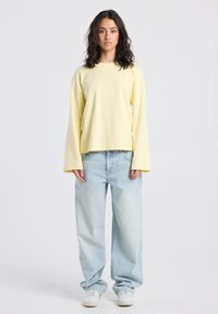 Young woman standing wearing a loose pale yellow long-sleeve top, light blue wide-leg jeans, and white sneakers on a plain background.