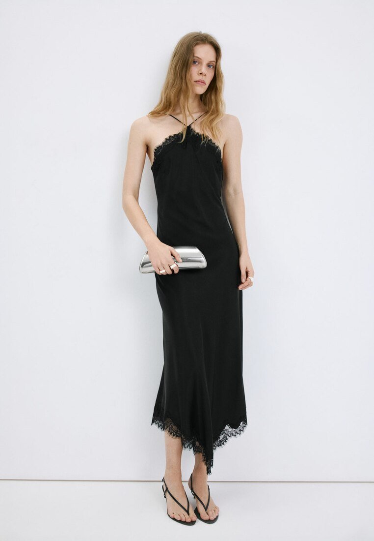 Femme portant une robe nuisette noire bordée de dentelle et des talons noirs à brides, tenant une pochette argentée sur un fond blanc uni.