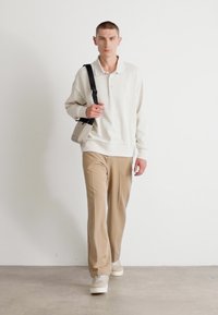 Weißer Sweater, beigefarbene Hose und helle Sneakers. Das Modell trägt eine hellgraue Schultertasche. Einfaches Design, lässige Passform, glatte Textur.
