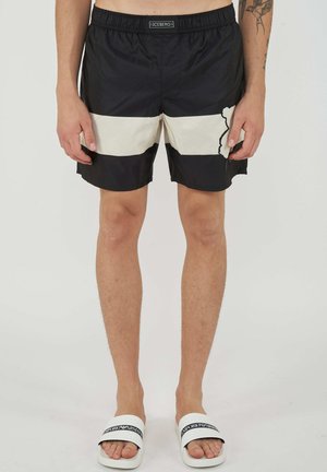 Personne portant un short de bain noir et beige à blocs de couleur avec taille élastique et des sandales slides blanches Emporio Armani.
