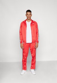 Survêtement rouge vif en matériau lisse, avec des rayures latérales bleues portant des logos. Associé à un t-shirt blanc et des baskets blanches.