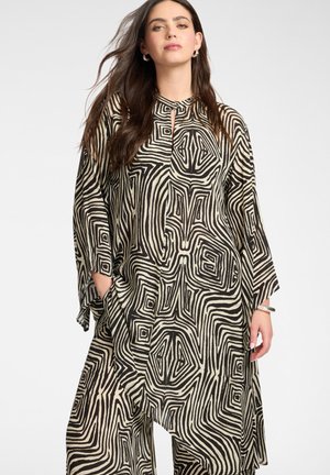 Kobieta z długimi ciemnymi włosami, ubrana w luźną sukienkę w abstrakcyjny czarno-beżowy wzór, pozująca z rękami w kieszeniach.