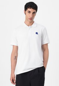 Biela polo košeľa so zapínaním na tri gombíky, krátkymi rukávmi a modrým vyšitým logom na ľavej strane hrudníka. Vyrobená z bavlny.