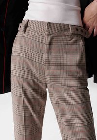 Gros plan sur une personne portant un pantalon à carreaux taille haute avec une ceinture et un haut côtelé blanc, se tenant devant un fond clair.