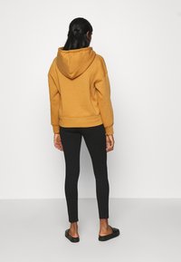 Mustard yellow hooded sweatshirt with a pocket, paired with black leggings. The texture appears soft and cozy, with a relaxed fit.