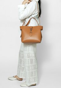 Borsa a spalla in cuoio tan con un'ampia apertura, un accento metallico circolare e una forma morbida e strutturata. Abbinata a pantaloni bianchi a gamba larga con texture.