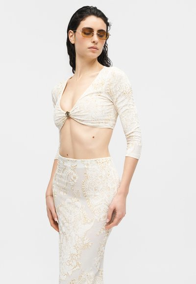 Γυναίκα που φοράει κρεμ και χρυσαφί μπλούζα με μοτίβο, με στριφτό μπροστινό μέρος και ταιριαστή ψηλόμεση φούστα, με γυαλιά ηλίου, στέκεται μπροστά σε ένα απλό φόντο.