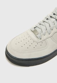 Light gray suede sneaker with perforated toe box, flat laces, and black rubber sole. Features a small branded accent near the laces.