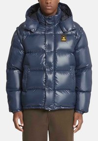 Giacca trapuntata blu navy con finitura lucida, design a quadri, collo alto e patch con logo. Presenta bottoni a pressione e tasche laterali.