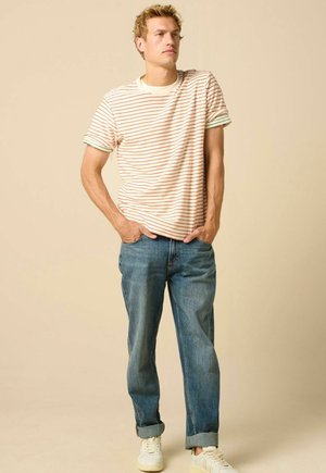 Junger Mann steht mit den Händen in den Taschen, trägt ein gestreiftes T-Shirt, blaue hochgekrempelte Jeans und weiße Sneakers vor einem einfarbigen Hintergrund.