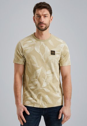 Camiseta beige claro con un patrón de hojas, mangas cortas, cuello redondo y una etiqueta negra en el pecho. Combinada con vaqueros azul oscuro.