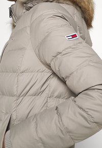 Beige prošívaná bunda s prošívaným designem, s kapucí podšitou kožešinou a emblémem se znakem na rukávu. Materiál vypadá hladce a izolačně.