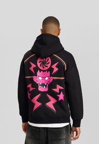 Man med svart huvtröja med ett klart rosa tecknat monsteransikte och blixtdesign på ryggen, stående mot en grå bakgrund.