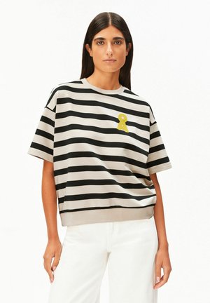 Vrouw met lang donker haar die een zwart-beige horizontaal gestreept T-shirt met korte mouwen draagt, voorzien van een klein geel lintspeldje, gecombineerd met een witte broek.