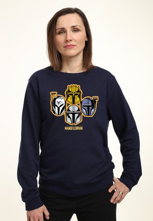 Donna che indossa una felpa blu navy con quattro elmi stilizzati Mandalorian e la scritta "Mandalorian" sul davanti.