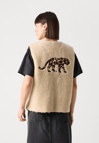 Beige Fleece-Weste mit einem Leopardenmuster-Applikation auf der Rückseite, abgerundeten Kanten und einer weichen Textur. Getragen über einem schwarzen Shirt.