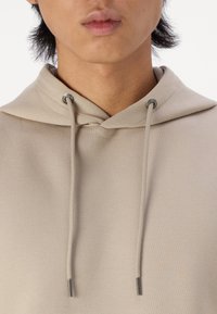 Bežový pulóver s kapucňou s textúrovaným povrchom, nastaviteľnými šnúrkami a striebornými kovovými prvkami. Voľný strih, minimalistický dizajn.
