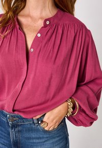 Blusa rosa con bottoni e dettagli plissettati sulle spalle. Indossata con jeans in denim blu e gioielli dorati al polso e sulle dita.
