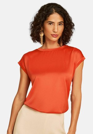 Kvinde med krøllet hår iført en lys orange kortærmet top og beige nederdel, accessoriseret med lange guldbøjler.