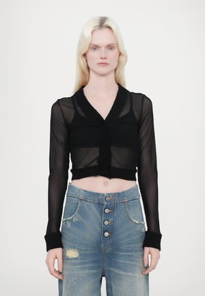 Gilet noir court en tissu en maille transparente, avec un col et des poignets côtelés texturés, porté sur un jean en denim bleu déchiré taille haute.