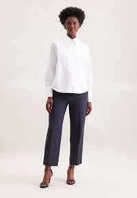 White button-up shirt with a rounded hem, paired with navy dress pants, worn with black strappy heels. Smooth fabric and fitted design.