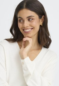 Pull blanc à col en V avec manches longues et détail de bouton sur le poignet. Le tissu semble doux et léger.