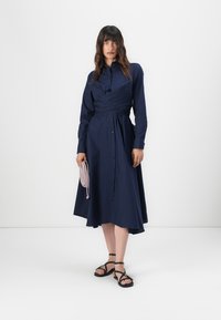 Marinblå skjortklänning med lång ärm, midjeintryck, knäppning fram och A-linjeformad kjol; ihop med en ljusrosa väska och svarta sandaler.