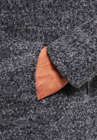 Abrigo de lana gris con texturas y un forro naranja llamativo en el bolsillo. La tela parece suave y esponjosa, con un diseño estructurado.