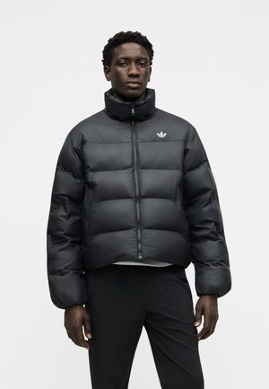 Junger Mann stehend, trägt eine schwarze Steppjacke mit hohem Kragen, zugezogen, und schwarze Hose vor hellem grauem Hintergrund.