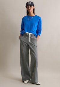 Niebieska bluza crewneck, szare spodnie o szerokich nogawkach z białym sznurkiem oraz niebieska czapka z daszkiem. Model stoi z rękami w kieszeniach.