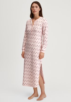 Vrouw die blootsvoets staat in een lange roze nachtjapon met een donker bloemenpatroon en een zijsplit, tegen een effen lichte achtergrond.