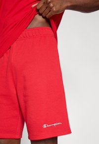 Κόκκινα κοντά παντελόνια από βαμβάκι με ελαστική μέση, που φέρουν το λευκό λογότυπο «Champion» στο αριστερό  πόδι. Το ύφασμα φαίνεται λείο και απαλό.