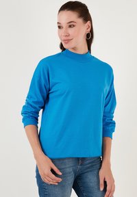 Sudadera azul con cuello alto, mangas largas y puños de canalé, hecha de tela suave. Se lleva con jeans de mezclilla de textura suave.