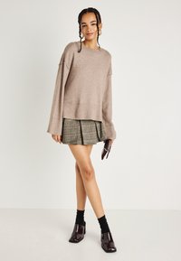 Μπεζ πλεκτό πουλόβερ σε oversized στυλ με φαρδιά μανίκια, συνδυασμένο με πράσινη καρό φούστα με πτυχώσεις. Το μοντέλο φοράει μαύρες κάλτσες και καφέ παπούτσια με τετράγωνο τακούνι.