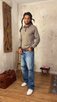 Homme avec des dreadlocks, pull beige, jean bleu et baskets blanches, debout dans une pièce rustique avec un sol en bois et une décoration murale.