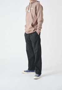Felpa marrone chiaro con logo bianco, pantaloni neri e sneakers in tela blu. La felpa presenta una tasca a marsupio e un cappuccio con cordino.