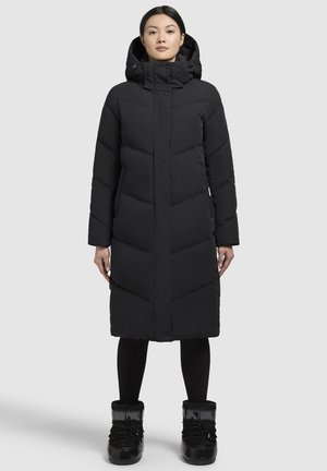 Zwarte pufferjas met een hoge kraag en capuchon, voorzien van een gewatteerd patroon, zijzakken en een verlengde snit. In combinatie met zwarte winterlaarzen.