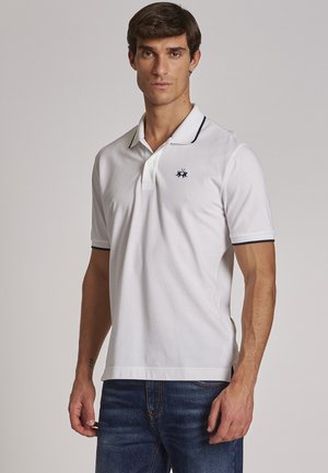 Hvid polo-shirt med marineblå detaljer, korte ærmer og en to-knaps placket. Har et broderet logo på brystet. Sammen med denim jeans.