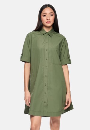 Young woman with long dark hair wearing a loose-fitting olive green button-up shirt dress with rolled-up sleeves against a plain background.