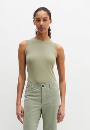 Femme aux cheveux bouclés portant un haut sans manches vert clair et un pantalon taille haute vert clair, debout devant un fond blanc uni.