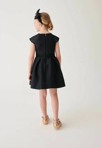 Schwarzes Kleid mit kurzen Ärmeln, schwingendem Rock und strukturiertem Taillenbund, kombiniert mit glänzenden bronzenen Ballerinas und einem schwarzen Haarband.