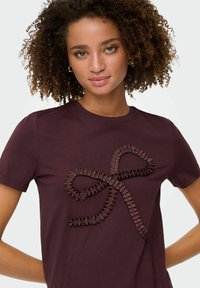 Młoda kobieta z kręconymi włosami, ubrana w bordowy t-shirt z teksturowanym wzorem w postaci wstążki na piersi, stojąca na tle białym.