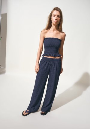 Mlada žena s dugom kosom nosi mornarskoplavu top bez naramenica s naborima, široke hlače u istoj boji i crne japanke na bijeloj pozadini.