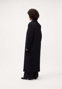 Lang sort frakke lavet af et tykt stof, med et skræddersyet design, store reverser og knaplukning på ærmerne, båret over brede sorte bukser.